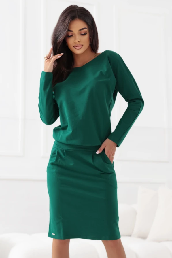 Modelka prezentuje elegancką zieloną sukienkę z długim rękawem i kieszeniami, podkreślającą kobiecą sylwetkę. Stylizacja łączy wygodę z klasycznym szykiem, idealna na co dzień i do pracy.