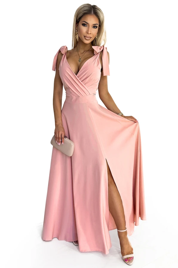 A long, elegant pink dress with a wrap-style bodice and a high slit, featuring delicate shoulder ties. The dress is complemented by a matching clutch and strappy heels, creating a sophisticated and stylish look.