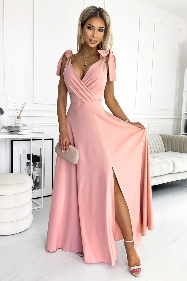 A long, elegant pink dress with a deep V-neckline and tied shoulder straps, featuring a flowing skirt with a high slit. The dress is complemented by a matching clutch and strappy heels, creating a sophisticated and stylish look.