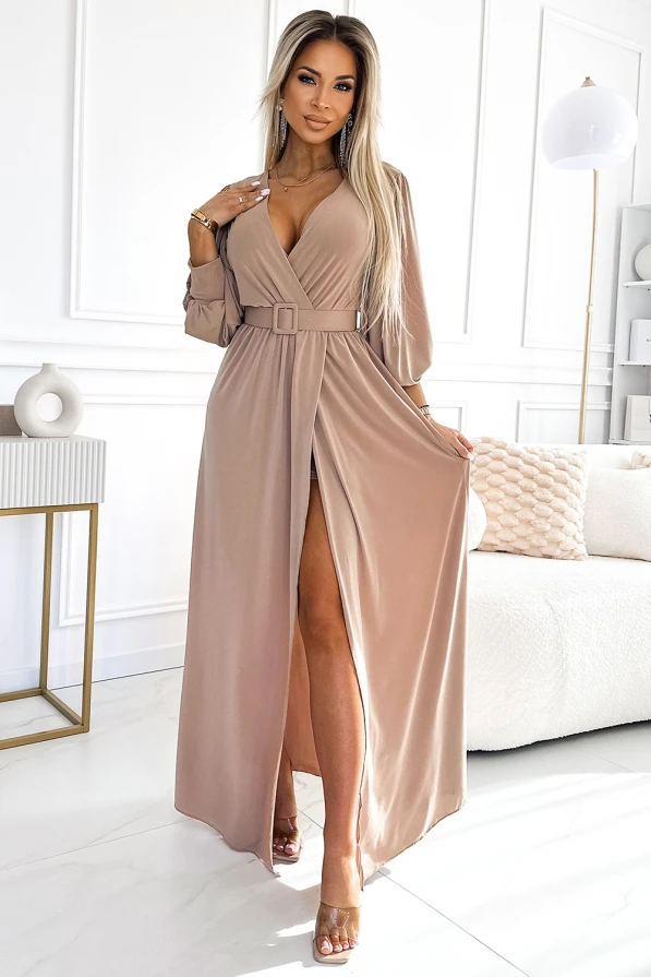 A long, elegant dress in a soft beige color features a deep V-neckline and a stylish belt at the waist, enhancing its sophisticated silhouette. The dress has a high slit, adding a touch of allure and modernity to its classic design.