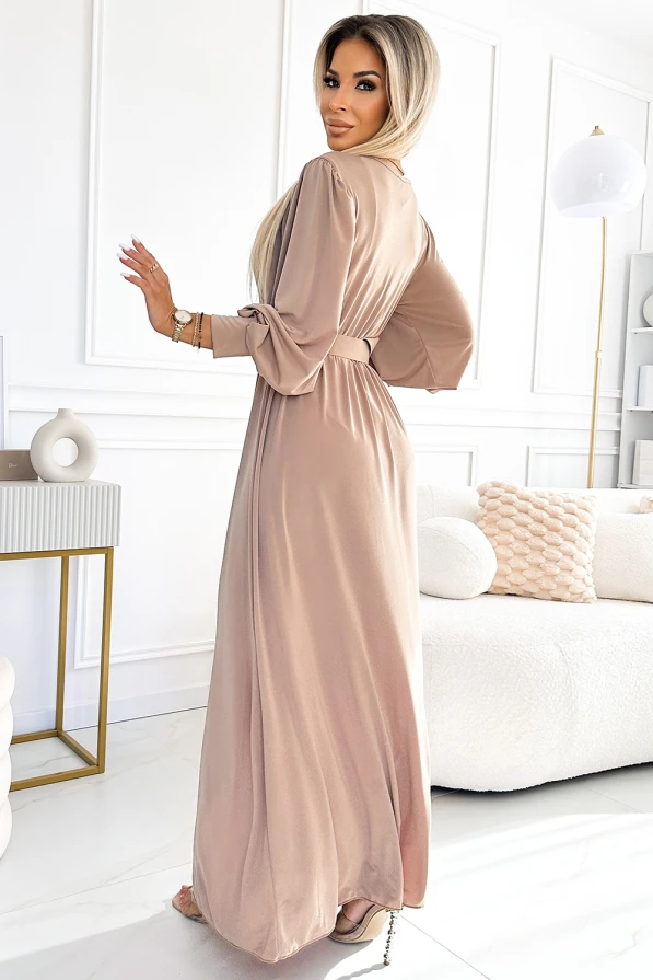 A long, elegant dress in a soft beige color features a flowing silhouette with a belted waist and stylish, loose sleeves. The fabric drapes gracefully, creating a sophisticated and timeless look perfect for formal occasions.