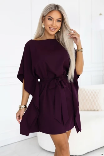 Model wearing a Numoco kimono dress in ripe plum, with a boat neckline, wide sleeves, a belt tied at the waist and draped sides, in a just-above-the-knee length.}
