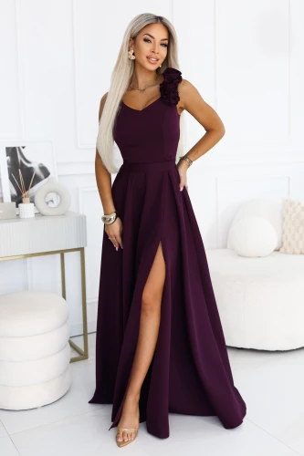 Model wearing a Numoco maxi dress in ripe plum, with a V-neckline, decorative roses on the shoulder, an accentuated waist and a flared bottom with a high leg slit.}