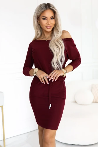 Model wearing a burgundy Numoco sporty dress in a just-above-the-knee length, with a boat neckline baring one shoulder, 3/4 sleeves, side pockets and a waist tie.}
