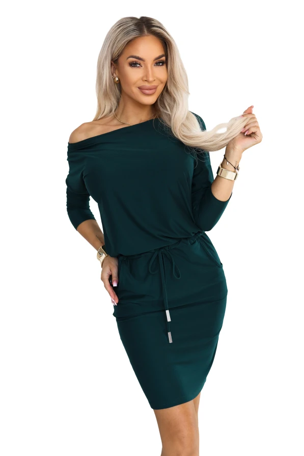 Model wearing a sporty dress in bottle green, with a neckline baring one shoulder, 3/4 sleeves, side pockets and a waist tie. A cut with a loose top and a fitted bottom just above the knee.