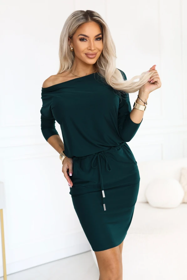 Model wearing a sporty dress in bottle green, with a neckline baring one shoulder, 3/4 sleeves, side pockets and a waist tie. A cut with a loose top and a fitted bottom just above the knee.