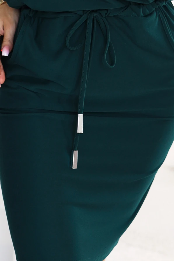 Model wearing a sporty dress in bottle green, with a neckline baring one shoulder, 3/4 sleeves, side pockets and a waist tie. A cut with a loose top and a fitted bottom just above the knee.