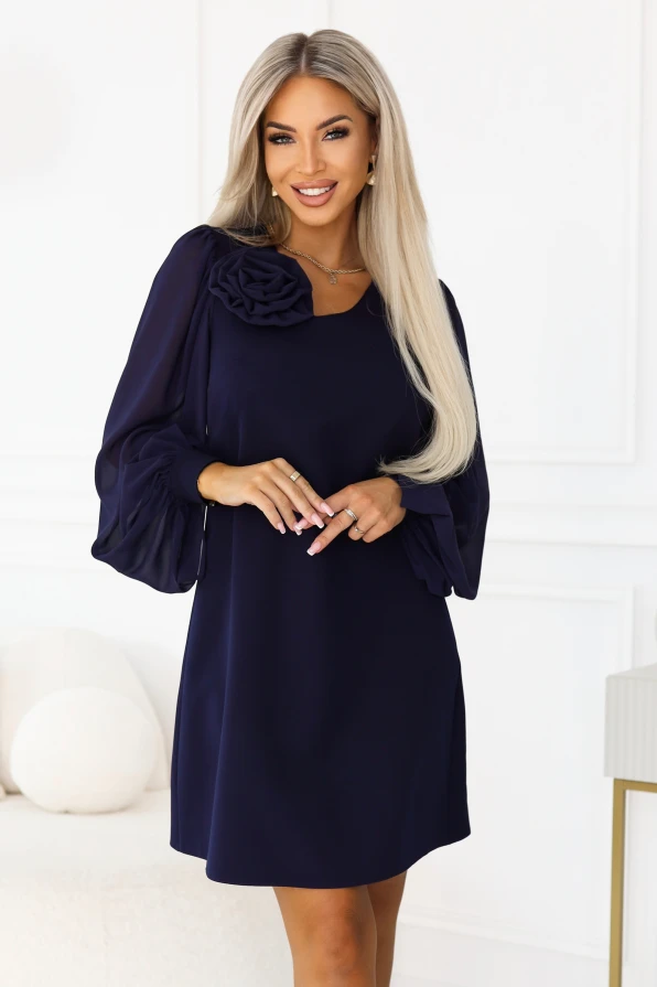 Model wearing the navy blue NELIA dress by Numoco in a trapeze cut just above the knee, with a decorative rose at the neckline and long, sheer sleeves finished with wide cuffs with a button.