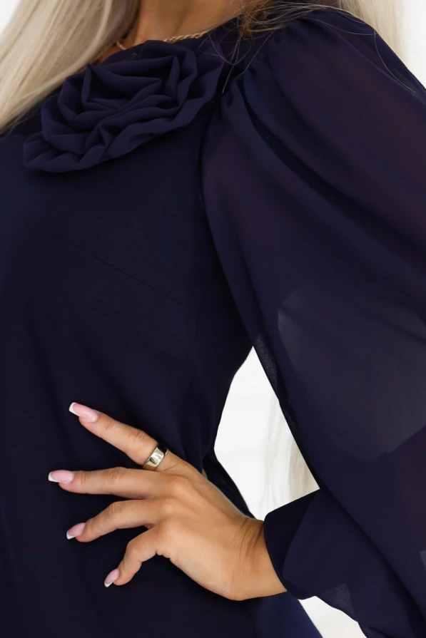 Model wearing the navy blue NELIA dress by Numoco in a trapeze cut just above the knee, with a decorative rose at the neckline and long, sheer sleeves finished with wide cuffs with a button.