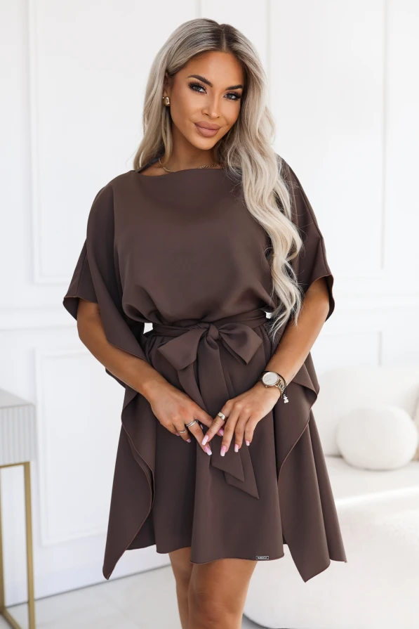 Model wearing the SOFIA dress by Numoco in a mocha brown shade. The dress reaches just above the knee, with a boat neckline, wide butterfly sleeves and a belt tied at the waist in a bow.