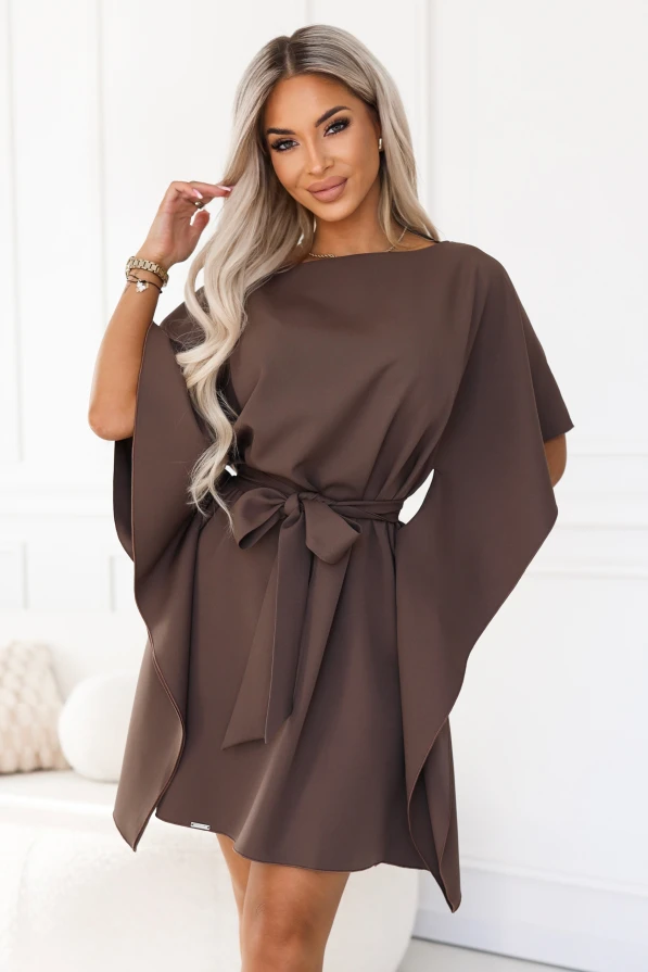 Model wearing the SOFIA dress by Numoco in a mocha brown shade. The dress reaches just above the knee, with a boat neckline, wide butterfly sleeves and a belt tied at the waist in a bow.