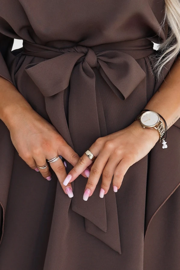 Model wearing the SOFIA dress by Numoco in a mocha brown shade. The dress reaches just above the knee, with a boat neckline, wide butterfly sleeves and a belt tied at the waist in a bow.